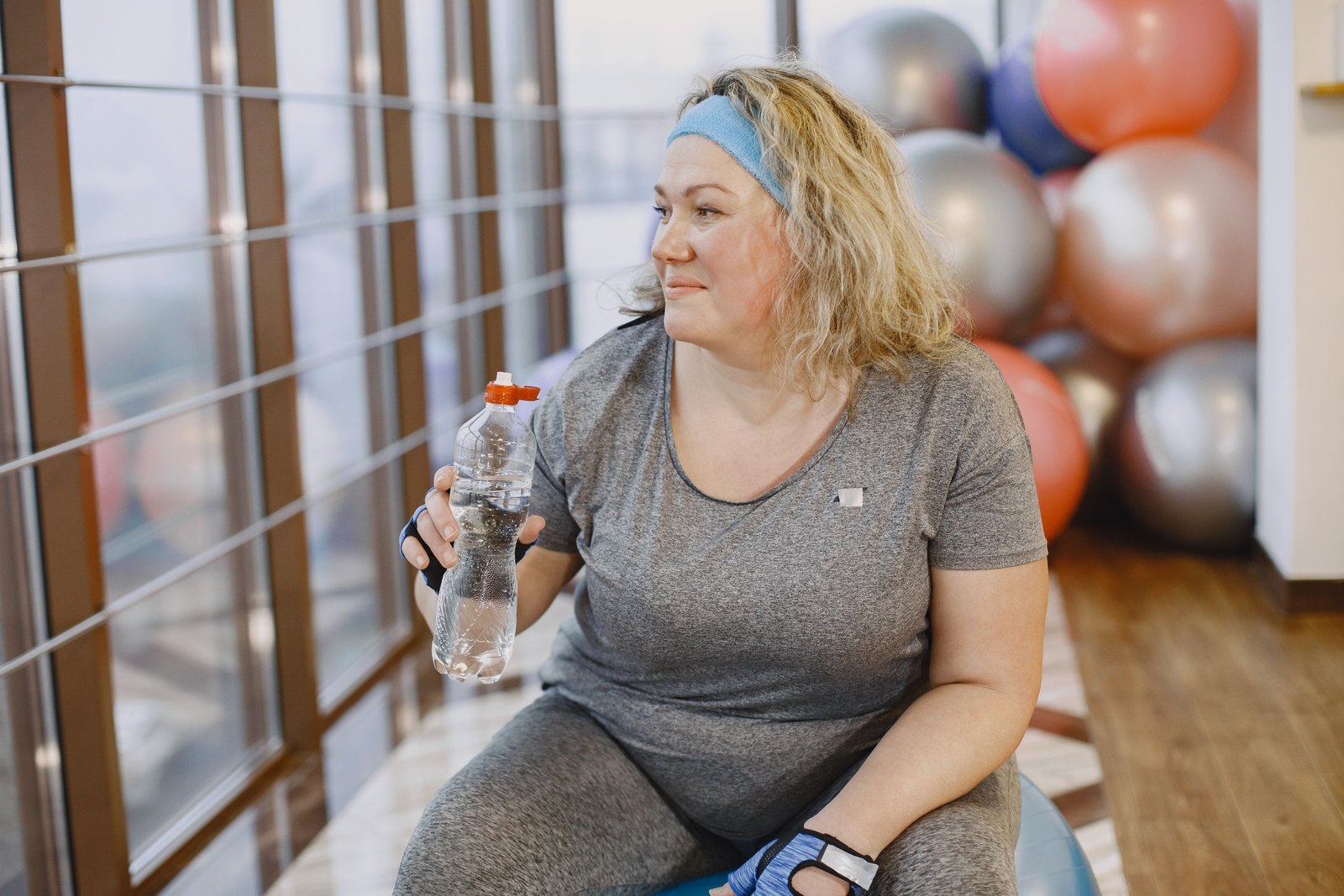 A woman taking a break on an exercise ball in a gym, water bottle in hand
