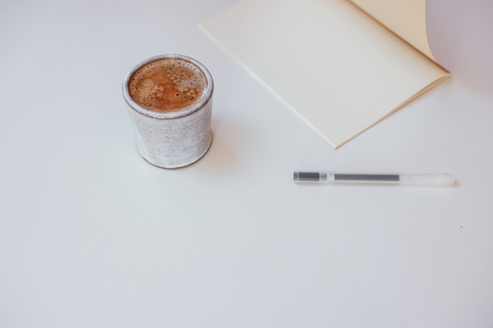 A cup of coffee beside a closed notebook and a pen on a white table