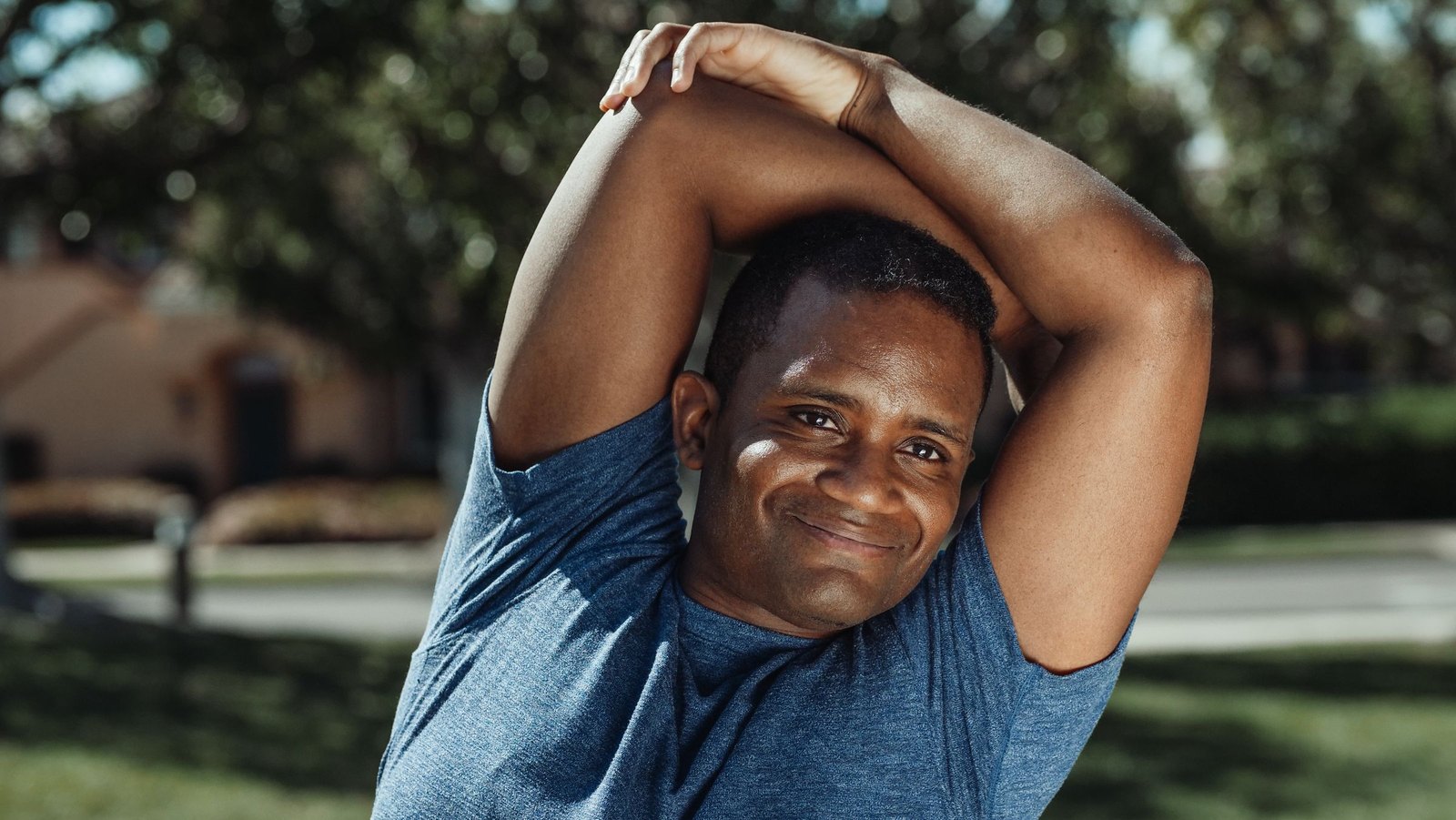 A man doing an overhead stretch outdoors, smiling at the camera