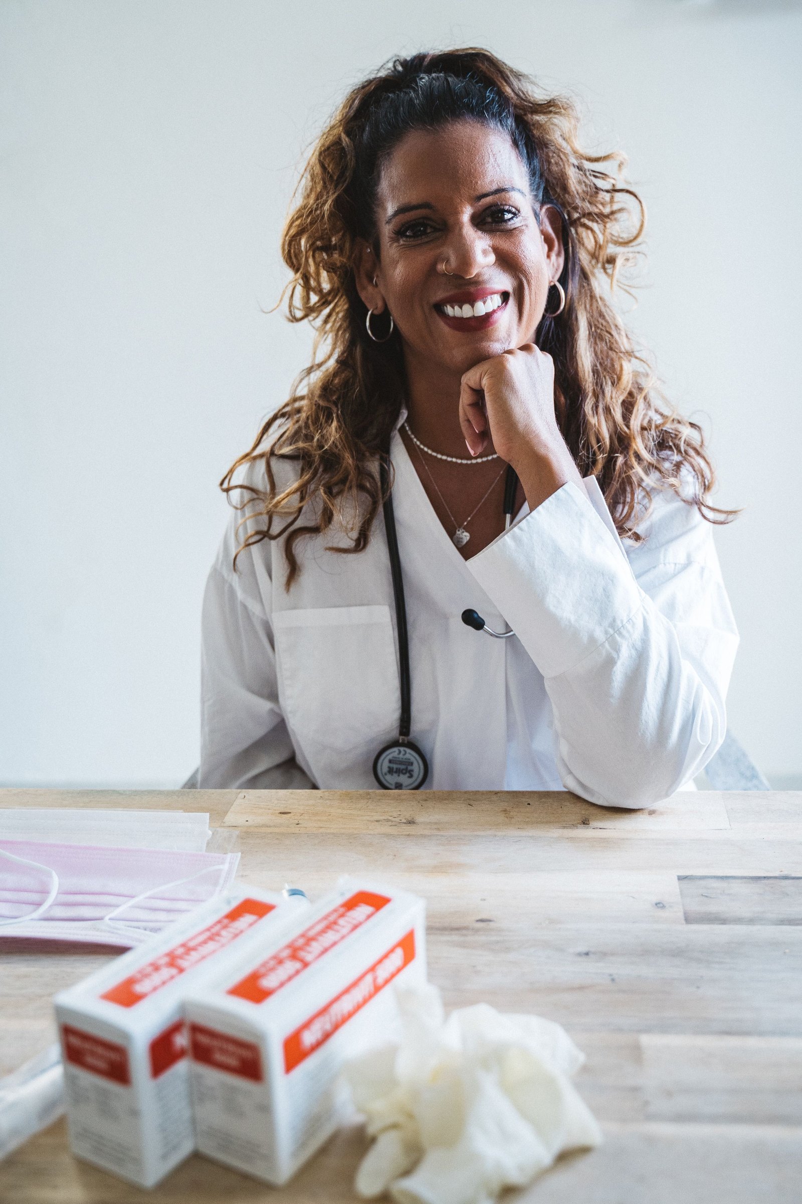 A doctor in a white coat seated at a desk, smiling with her chin on her hand